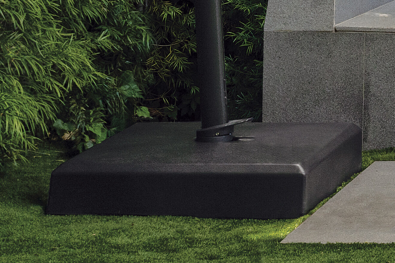 Outdoor view of the AKZ Sand-Filled Resin Base in black finish positioned on lush green artificial turf beside a grey stone wall and paving slabs. The sturdy, flat-topped base supports a black cantilever umbrella pole fitted through its central mounting hole, secured by a dark metal bracket and locking foot pedal. The base’s broad square profile and matte surface emphasise its durable construction and balanced weight distribution for anchoring large umbrellas.