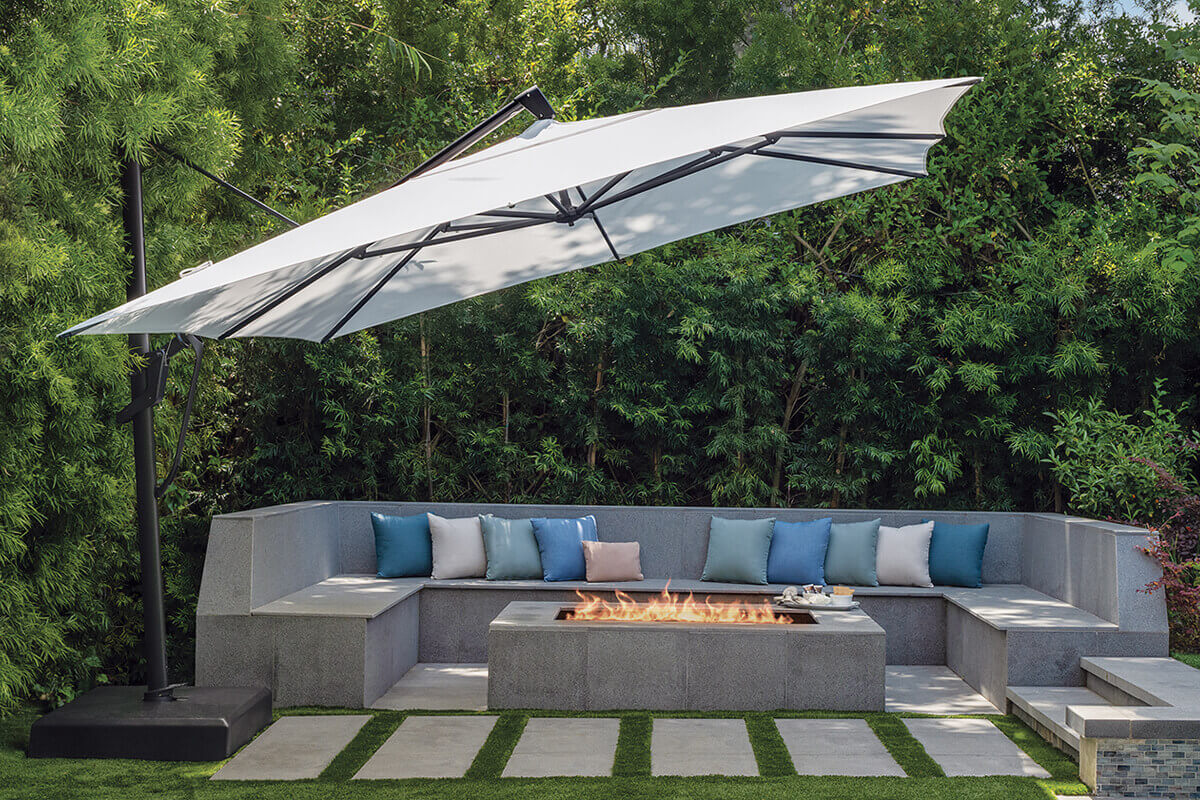 Outdoor setting showing a large AKZ Plus cantilever umbrella with a light grey canopy shading a modern concrete lounge area. The umbrella is positioned behind an L-shaped sectional bench with multicoloured blue and grey cushions surrounding a rectangular fire feature. The umbrella’s sturdy black frame and side post are secured to a sand-filled resin base on manicured grass tiles, with lush green bamboo foliage forming a private backdrop.