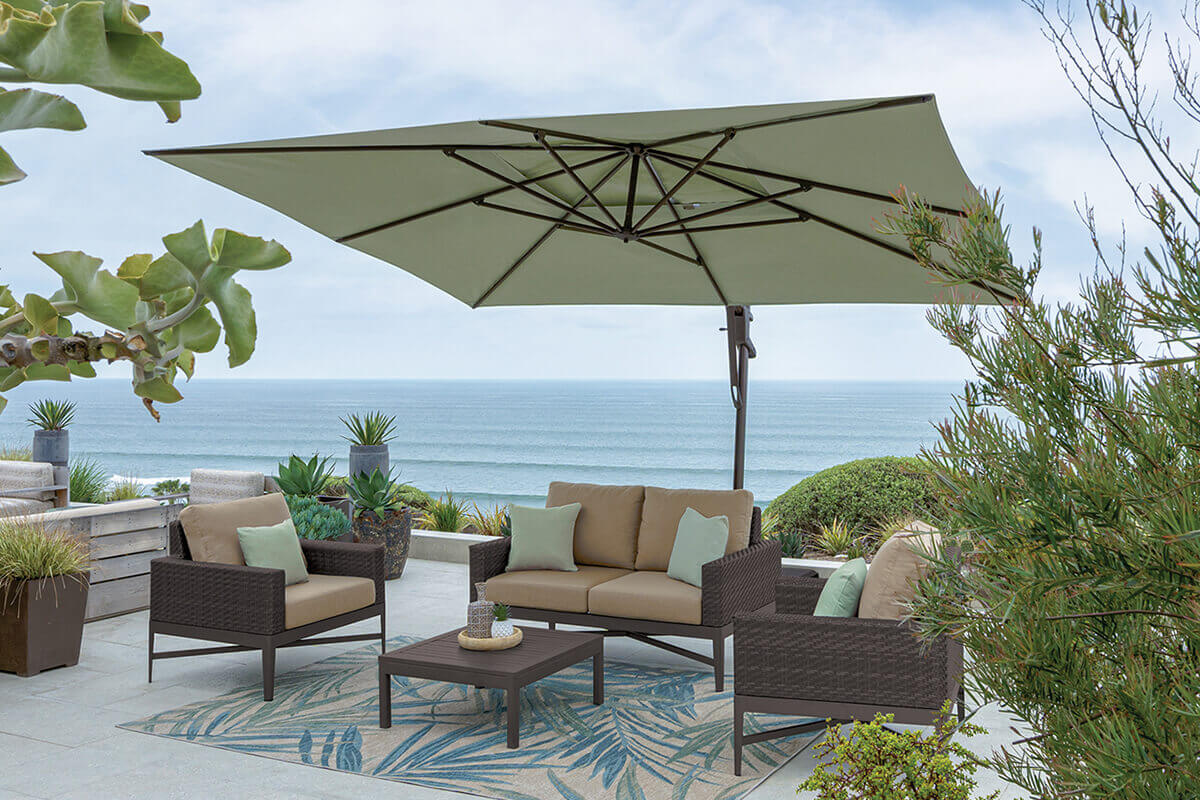 Beachfront patio scene featuring a tan and dark brown wicker lounge set shaded by a sage green AKZ Plus cantilever umbrella. The umbrella’s canopy is extended over a coffee table with potted plants and a patterned rug beneath, while ocean waves roll in the background under a bright, slightly overcast sky. The side pole and base sit neatly on the stone patio, complementing the coastal design.