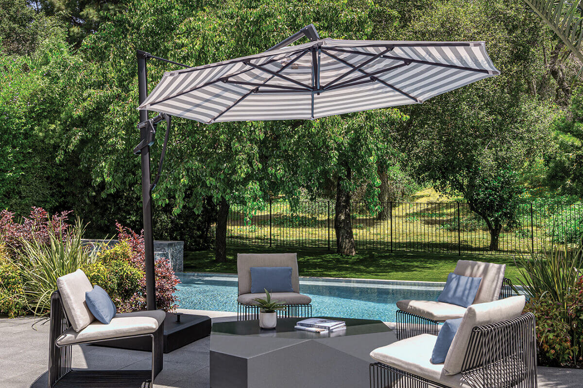Poolside outdoor area showcasing a black and white striped AKZ Plus cantilever umbrella fully extended over a modern seating arrangement. Four low chairs with beige cushions and blue accent pillows surround a dark geometric coffee table on a tiled patio. The umbrella’s black frame and robust base blend with the sleek contemporary aesthetic, while trees, greenery, and a shimmering pool create a relaxed, resort-like atmosphere.