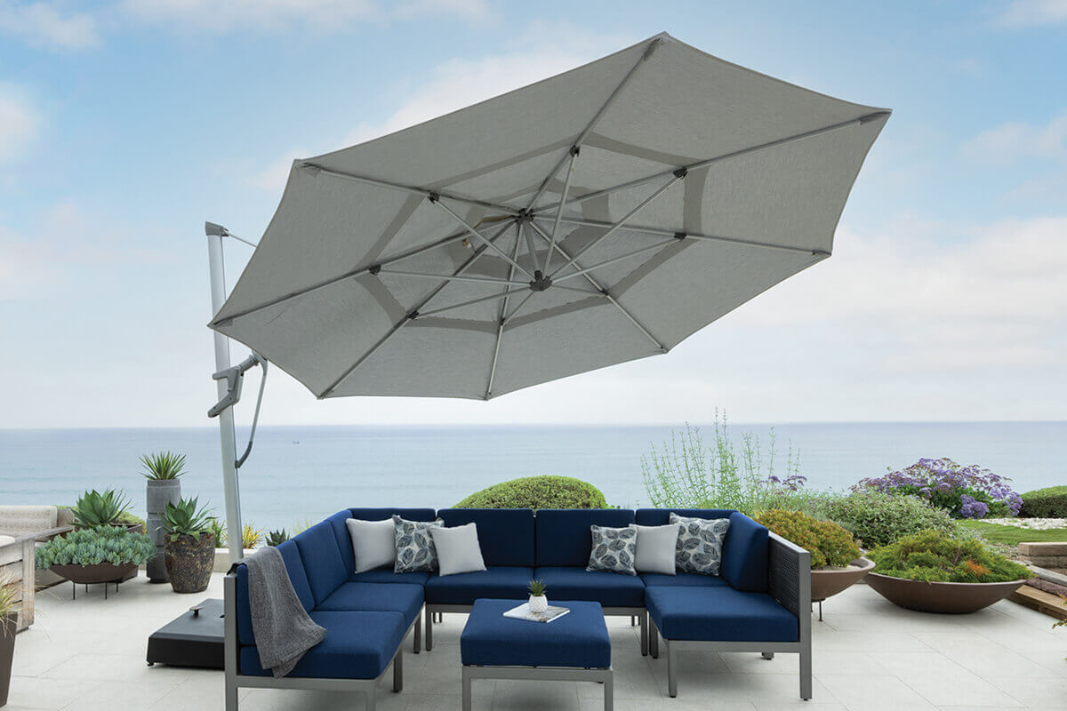 Rooftop terrace view of a grey AKZ Plus cantilever umbrella positioned beside a blue modular sectional sofa set overlooking the ocean. The umbrella’s octagonal canopy is open wide, supported by a silver aluminium frame and pole mounted on a rectangular resin base. The setting includes coastal plants, white stone flooring, and accent cushions, presenting an elegant, breezy coastal aesthetic ideal for outdoor entertaining.