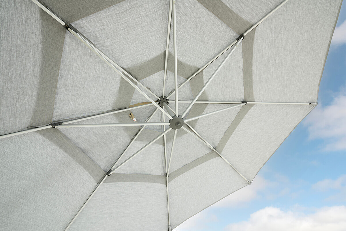 Close-up view from beneath the grey AKZ Plus umbrella canopy, showing its symmetrical eight-rib aluminium frame and robust hub mechanism. The light filters through the textured fabric panels, highlighting the durable construction and clean stitching. A small manufacturer’s label is visible on one of the inner seams, reinforcing attention to design detail and quality craftsmanship.