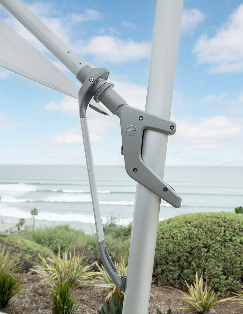 Detailed close-up of the AKZ Plus umbrella’s tilt and locking mechanism, featuring the sleek light grey handle and rotation joint fixed to the aluminium mast. The background shows ocean waves and coastal vegetation, illustrating the umbrella’s versatility and modern engineering designed for effortless adjustment and 360-degree rotation.