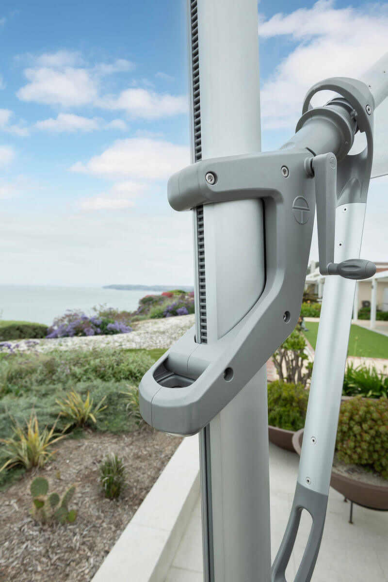 Close-up of the AKZ Plus umbrella’s sliding pistol grip handle and adjustment system on its vertical mast. The precision-moulded grey handle includes ergonomic contours and visible internal tracks for smooth gliding. Behind it, a coastal landscape with greenery and homes underscores its stylish integration into upscale outdoor environments.