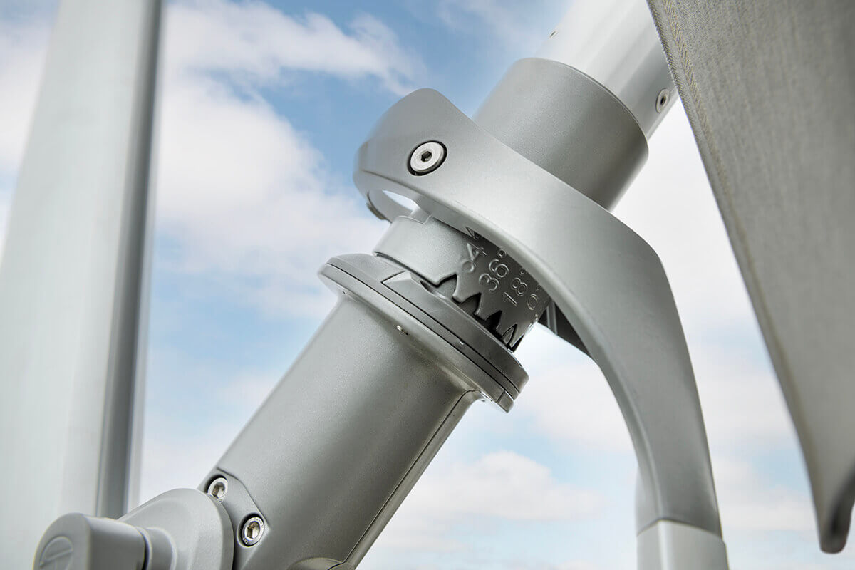 Macro view of the AKZ Plus umbrella’s silver rotation gear joint displaying engraved degree markings for angle adjustment. The gear-like teeth interlock securely within the pivoting mechanism, ensuring controlled tilting. The silver components contrast against the soft blue sky, highlighting the umbrella’s refined mechanical engineering.