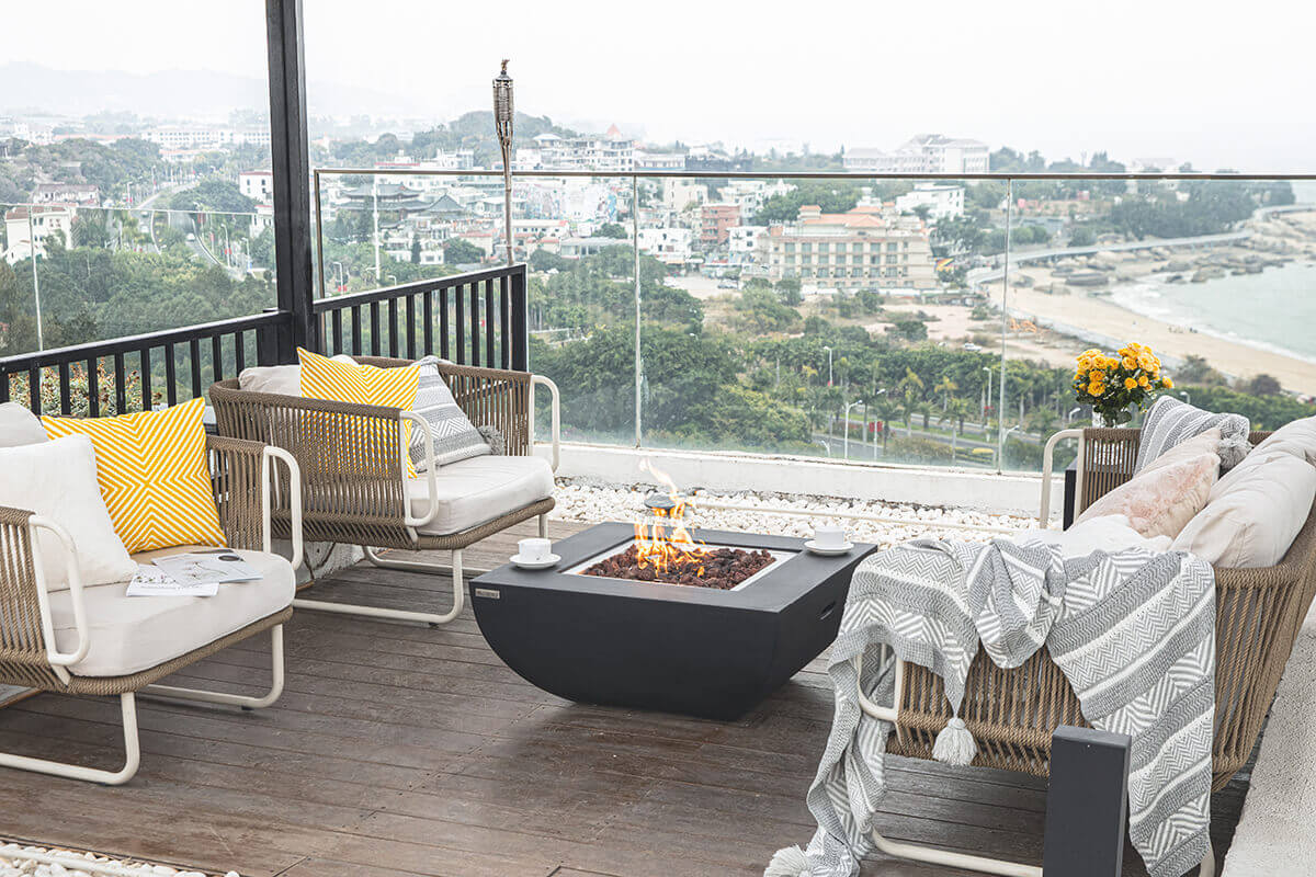 A rooftop patio styled with neutral outdoor lounge furniture arranged around a black Modeno Aurora fire table, which features a smooth glass fiber reinforced concrete body. The square fire table sits at the centre of the scene with its recessed burner filled with dark lava rock, producing a tall, bright flame. Two white teacups rest on the tabletop edges. The surrounding seating includes woven rope armchairs with thick cream cushions and yellow patterned throw pillows. A grey textured blanket drapes over one chair on the right. A glass safety railing frames the elevated patio and reveals a distant coastal city landscape with trees, buildings, shoreline and a hazy sky.