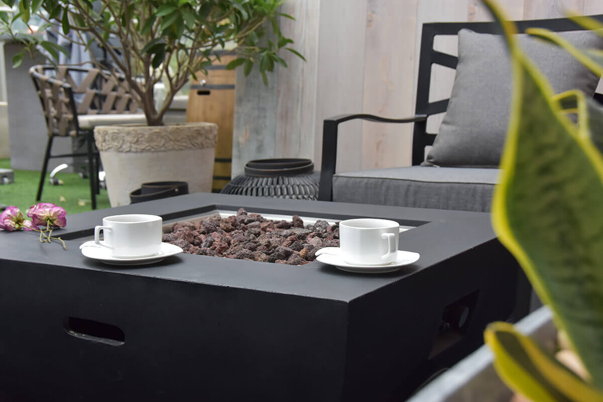 A close up view of the Modeno Aurora fire table showing two white teacups positioned near the front edge of the smooth black tabletop. The recessed square burner filled with dark textured lava rock is centred in the frame. The background reveals part of a grey cushioned lounge chair and indoor greenery including a decorative potted tree. The image highlights the table’s sharp edges, clean lines and fine concrete surface texture.
