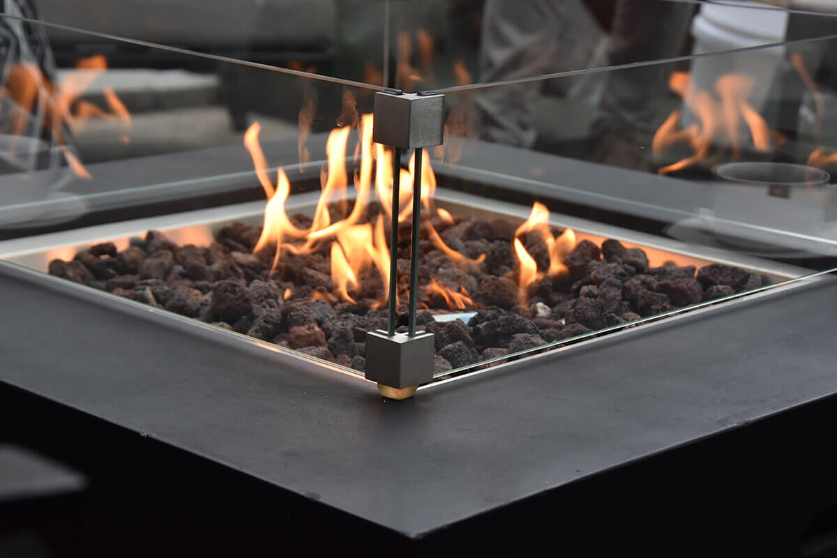 A detailed close view of the Aurora fire table’s flame display with vibrant orange and yellow flames rising through a bed of irregularly shaped lava rocks. A clear glass windscreen surrounds the burner, supported at each corner by stainless steel brackets with brushed metal surfaces. The black concrete tabletop fills the lower portion of the frame, showcasing its smooth matte finish.