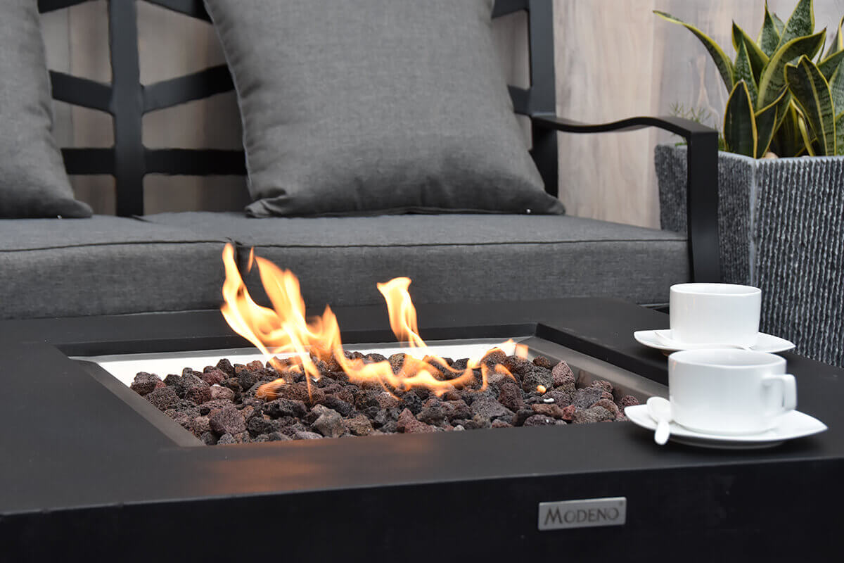 A close up shot of the Aurora fire table showing the active flame above dark lava rock while focusing on the smooth black tabletop surface and the Modeno brand badge mounted on one corner. Two white teacups with matching saucers sit near the right edge. A grey cushioned seat is visible in the background with a textured concrete planter positioned to the right.