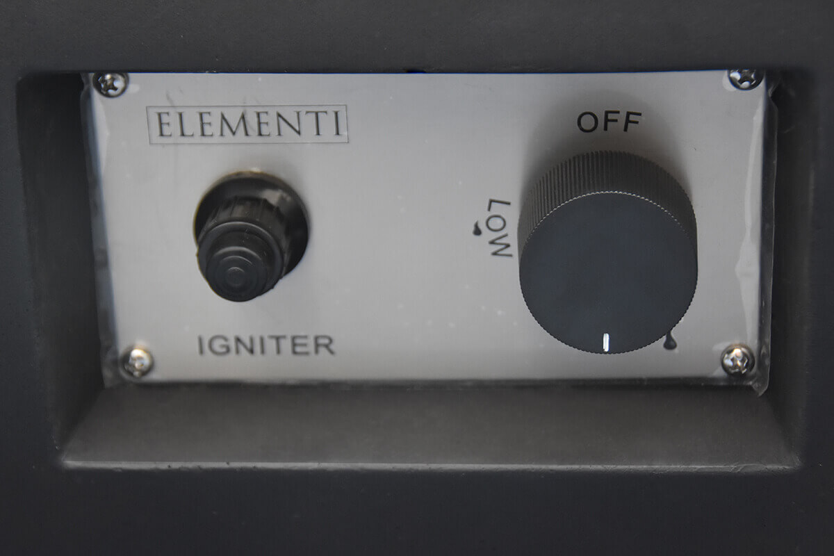 A close up view of the Elementi brand igniter control panel installed on the body of the Aurora fire table. The panel is made of brushed metal with the Elementi logo engraved at the top. On the left is a raised round push button igniter surrounded by a black housing. On the right is a textured circular control knob marked with Low and Off settings. Four visible screws secure the panel to the fire table’s concrete structure.