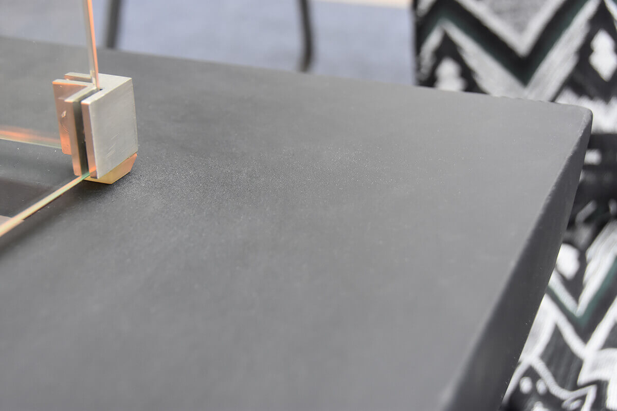 A macro level close up of the smooth black surface of the Aurora fire table’s glass fiber reinforced concrete top. The texture shows subtle matte grain and soft surface variations. The lower left corner includes a stainless steel corner bracket from the optional glass windscreen resting on the tabletop while a patterned blanket in black and grey appears blurred in the background.