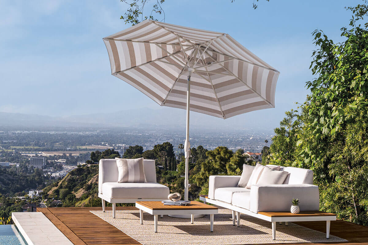 Beige and white striped Auto-Tilt Market Umbrella shading a white modular outdoor sofa set on a wooden deck high above a city. The umbrella’s striped canopy is fully extended on a white aluminium pole, casting cool shade over low-profile cushions while a sweeping valley, distant buildings, and hazy mountains fill the background.