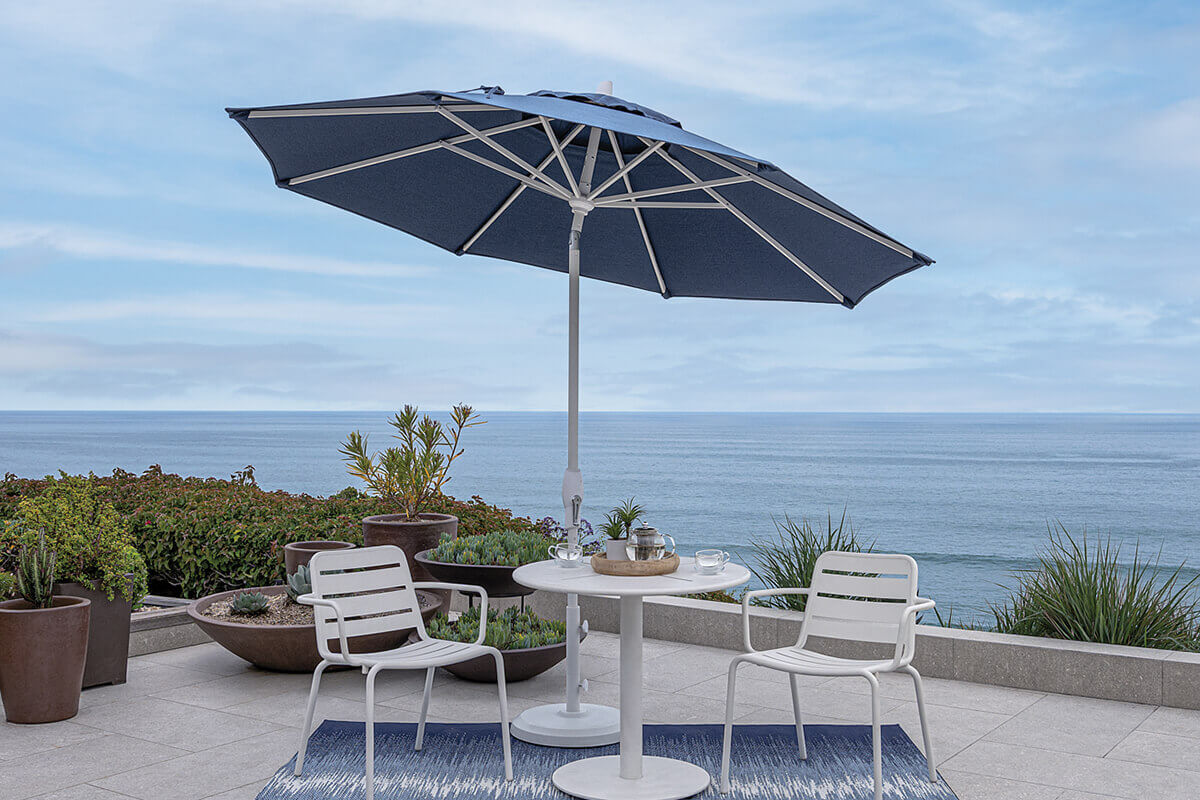 Navy blue Auto-Tilt Market Umbrella with a deep-coloured canopy open over a small white round café table and two white metal chairs on a tiled terrace. The umbrella’s white pole and ribs frame the dark fabric, while lush potted plants and a wide ocean horizon create a calm seaside setting.