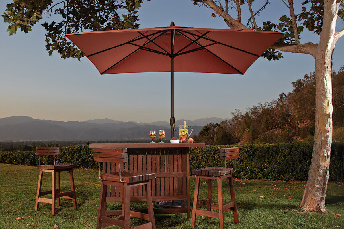 Terracotta Auto-Tilt Market Umbrella shading a wooden outdoor bar table and four matching bar stools on a manicured grass lawn. The rich orange canopy is fully open on a bronze pole, providing an intimate shaded spot for colourful drinks and fruit, with neatly trimmed hedges, distant hills, and a warm evening sky in the background.