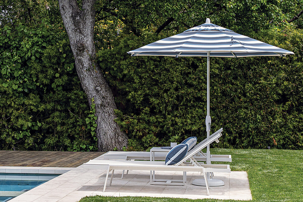 Blue-and-white striped Auto-Tilt Market Umbrella positioned beside a rectangular swimming pool, shading two white cushioned sun loungers. The striped canopy curves over the loungers from a white pole anchored to a round base on the pale stone deck, while dense green shrubs and a large tree trunk create a private, relaxing poolside retreat.
