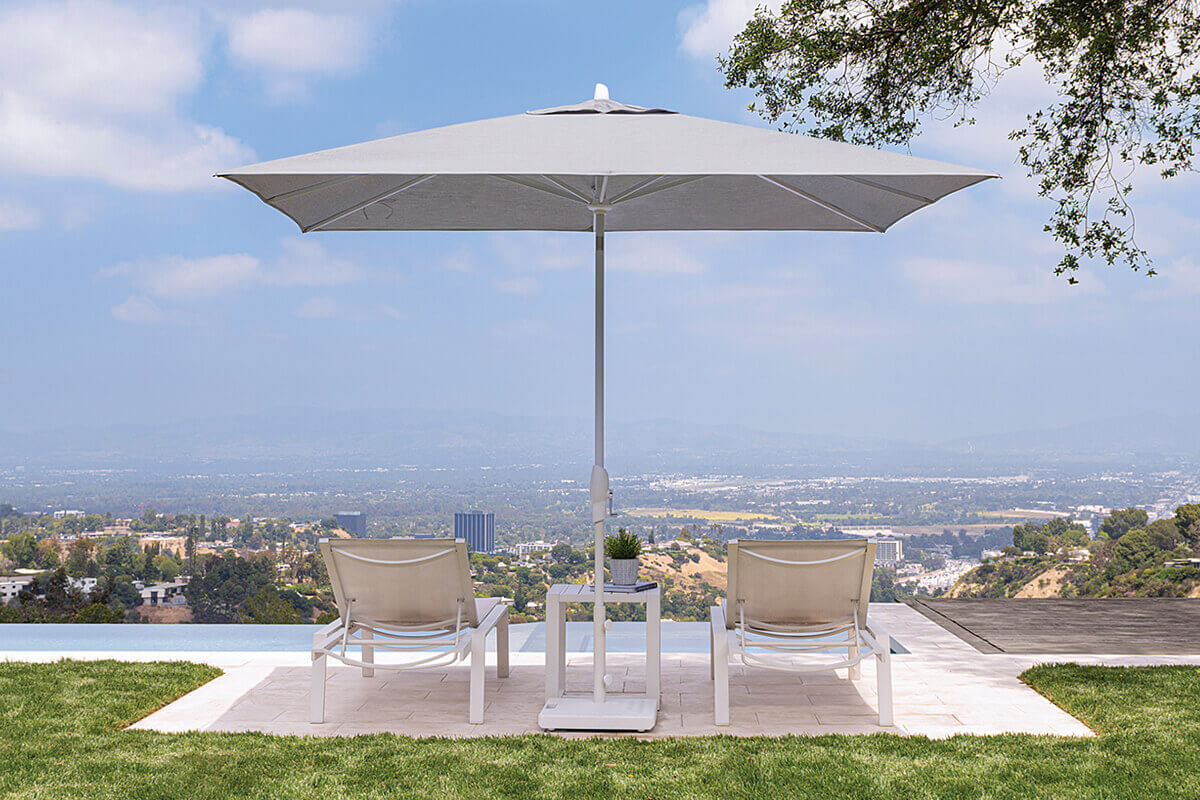 Light grey Auto-Tilt Market Umbrella standing behind two white chaise loungers at the edge of an infinity pool. The wide square canopy stretches evenly over the loungers, with a small white side table and potted plant between them. Beyond the pool, a broad valley dotted with buildings and hills fades into a soft blue sky filled with light clouds.