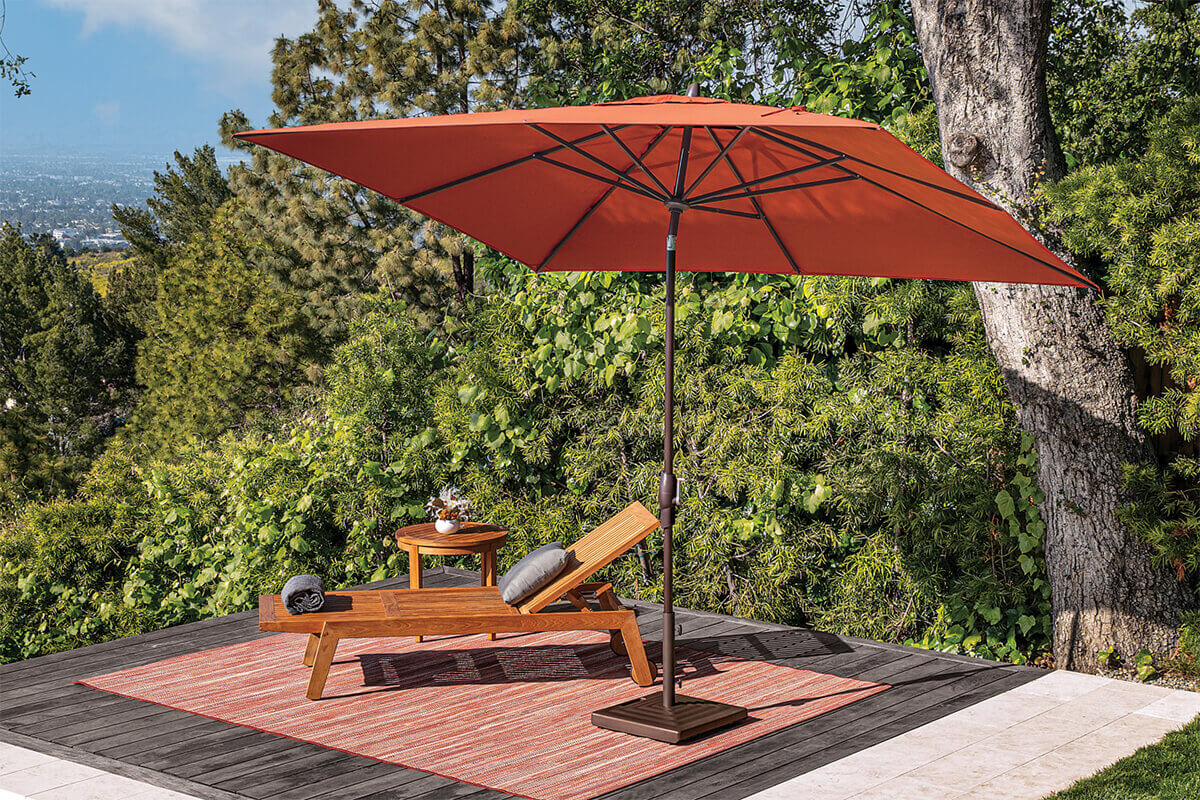 Burnt orange Auto-Tilt Market Umbrella shading a single wooden lounger with grey cushions on a dark deck platform. A patterned outdoor rug extends under the lounger, while the square canopy opens from a bronze pole anchored in a matching square base. Tall evergreens and lush foliage surround the deck, creating a secluded garden escape.