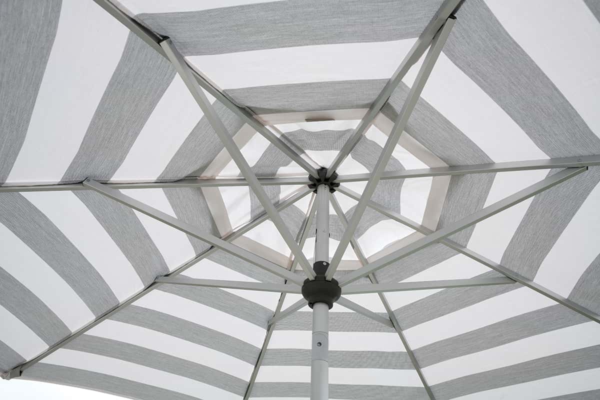 Under-canopy view of a grey-and-white striped Auto-Tilt Market Umbrella showing the internal aluminium rib system. The slim, pale ribs radiate from a central hub beneath alternating wide grey and white fabric panels, and the vented centre section reveals soft daylight, highlighting the even tension and sturdy construction of the canopy.