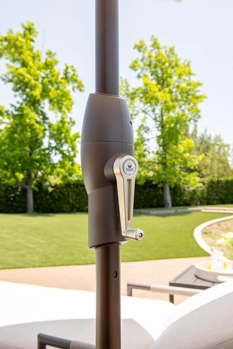 Close-up of the Auto-Tilt Market Umbrella crank mechanism mounted on a bronze-coloured aluminium pole. The rounded housing holds an ergonomic silver crank handle engraved with the Treasure Garden logo. Behind the pole, a sunlit lawn and blurred trees create a soft green backdrop that emphasises the umbrella’s durable outdoor hardware.