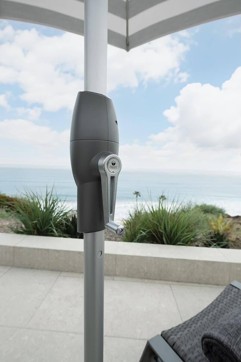 Silver crank handle on a grey Auto-Tilt Market Umbrella pole set against a coastal landscape. The sleek mechanism is fixed to the pole mid-height for easy reach, with the canopy edge and sky just visible at the top of the frame. In the distance, layered ocean waves, dunes, and a light blue sky with scattered clouds reinforce the umbrella’s seaside-ready design.