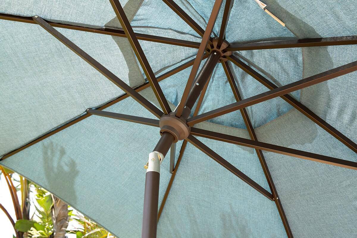 Underneath view of an Auto-Tilt Market Umbrella with a textured blue canopy supported by a brown aluminium centre pole and a circular hub. Multiple slim ribs extend outwards in a tidy radial pattern. Sunlight filters through the fabric, and the soft shadow of palm fronds appears on the canopy surface, suggesting a sunny poolside environment.