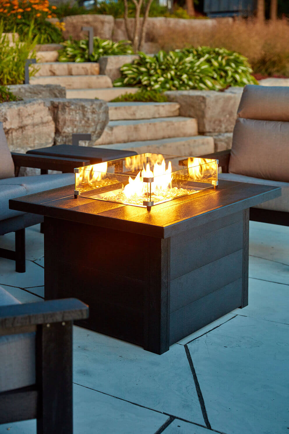 A rectangular outdoor fire table made from dark grey recycled plastic lumber sitting on a large stone patio beside a landscaped garden with stepped rock walls and lush green plants. The table surface displays a clear rectangular wind guard that surrounds a bed of clear fire glass with bright yellow flames rising from the centre. Two grey cushioned lounge chairs flank the table, creating a relaxed seating arrangement. Warm evening light reflects off the textured tabletop, highlighting its durable weatherproof construction and marine grade stainless steel hardware.