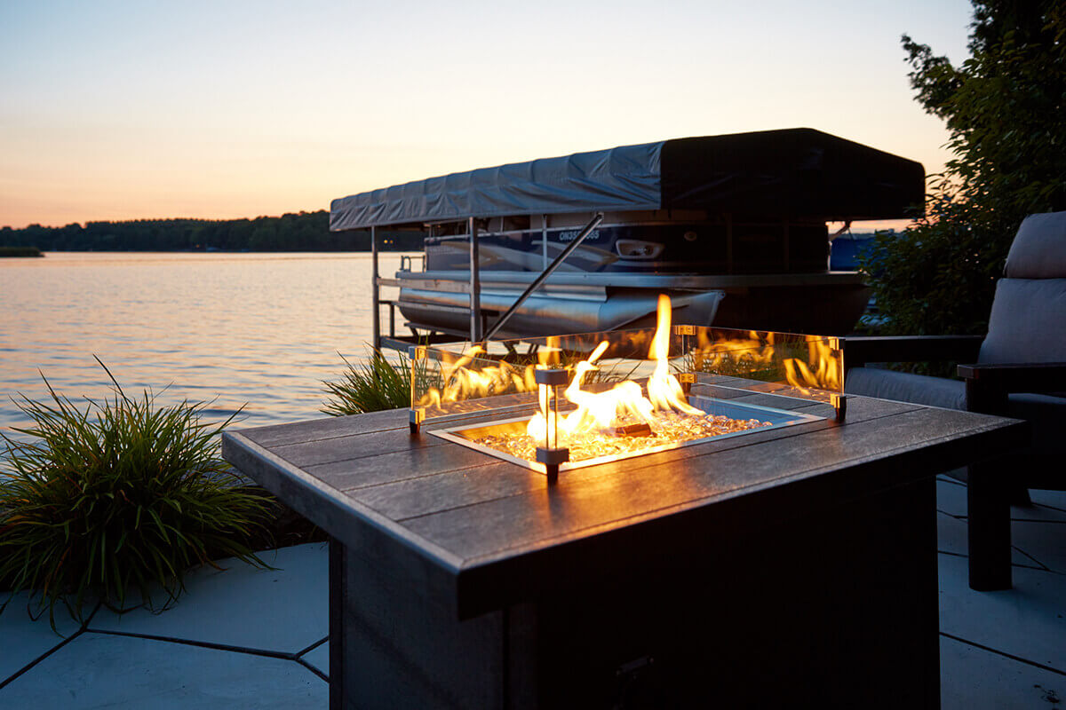 A view of the CRP rectangular fire table at sunset beside a calm lakeside dock. Yellow and orange flames flicker behind the clear wind guard, illuminating the grey recycled plastic tabletop with a warm glow. Decorative plants frame the scene while a covered pontoon boat floats nearby. The table sits between grey cushioned outdoor seating, creating a serene waterfront gathering space powered by the 12 by 24 Crystal Fire Plus burner.