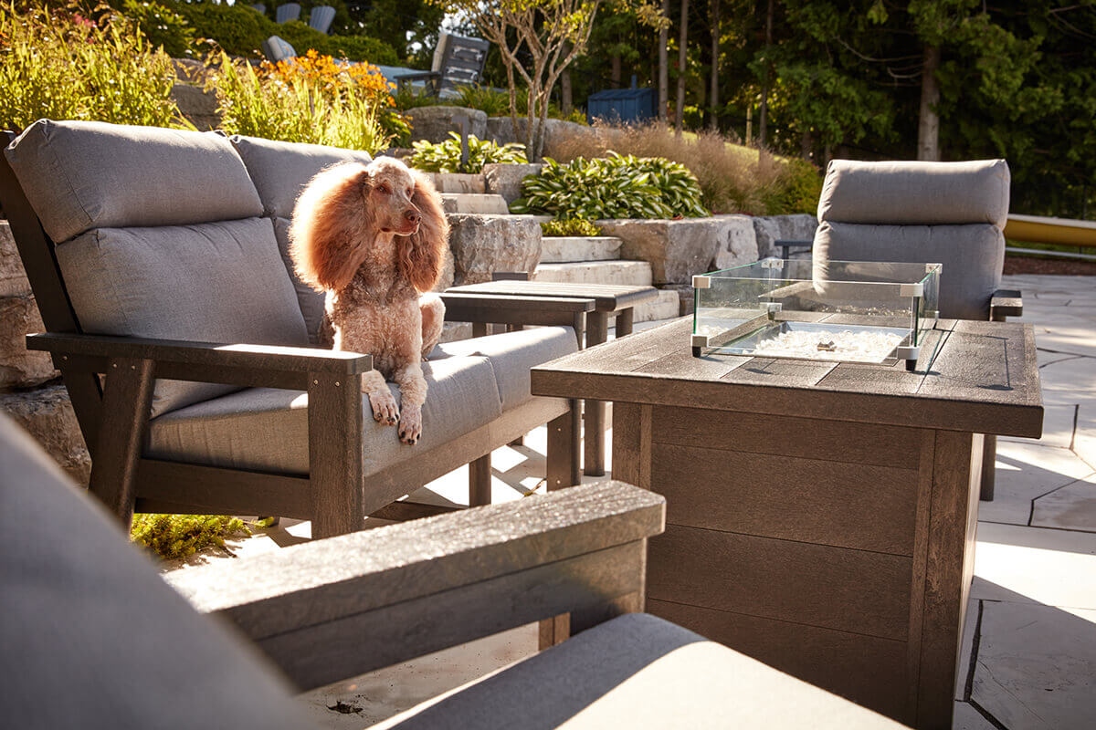A daylight scene showing the CRP rectangular fire table unlit, with the wind guard surrounding clear fire glass. A poodle with fluffy apricot coloured fur sits comfortably on a grey cushioned outdoor bench beside the table. Natural stone steps and lush landscaping rise in the background, emphasising the durability and weather resistance of the recycled plastic fire table positioned on the stone patio.