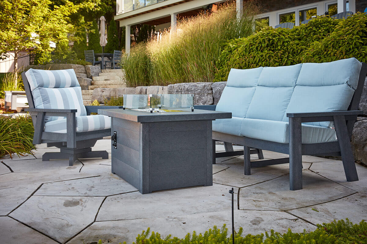 A lakeside patio setting with pale blue cushioned seating arranged around the CRP rectangular fire table. The unlit burner and clear wind guard sit at the centre of the spacious grey tabletop made from long lasting recycled plastic materials. Tall grasses, natural rock walls and a contemporary cottage exterior complete the elevated outdoor retreat.