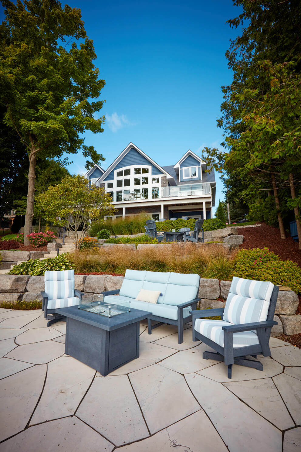 A wide view of a cottage property overlooking a large stone patio where the CRP rectangular fire table sits centred between pale blue cushioned outdoor seating. The unlit burner and clear wind guard rest on the dark grey tabletop. Towering trees, manicured landscaping and the large cottage with a wall of windows rise in the background, showcasing the fire table as a premium feature for relaxation and entertaining.
