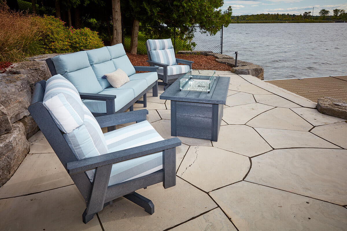 A lakeside seating area featuring the CRP rectangular fire table surrounded by pale blue and white striped cushioned chairs. The clear wind guard rests above the unlit burner. The table sits on a patterned stone patio next to the water while large rocks, evergreens and shrubs line the edge. The durable recycled plastic table blends seamlessly with the natural landscape.