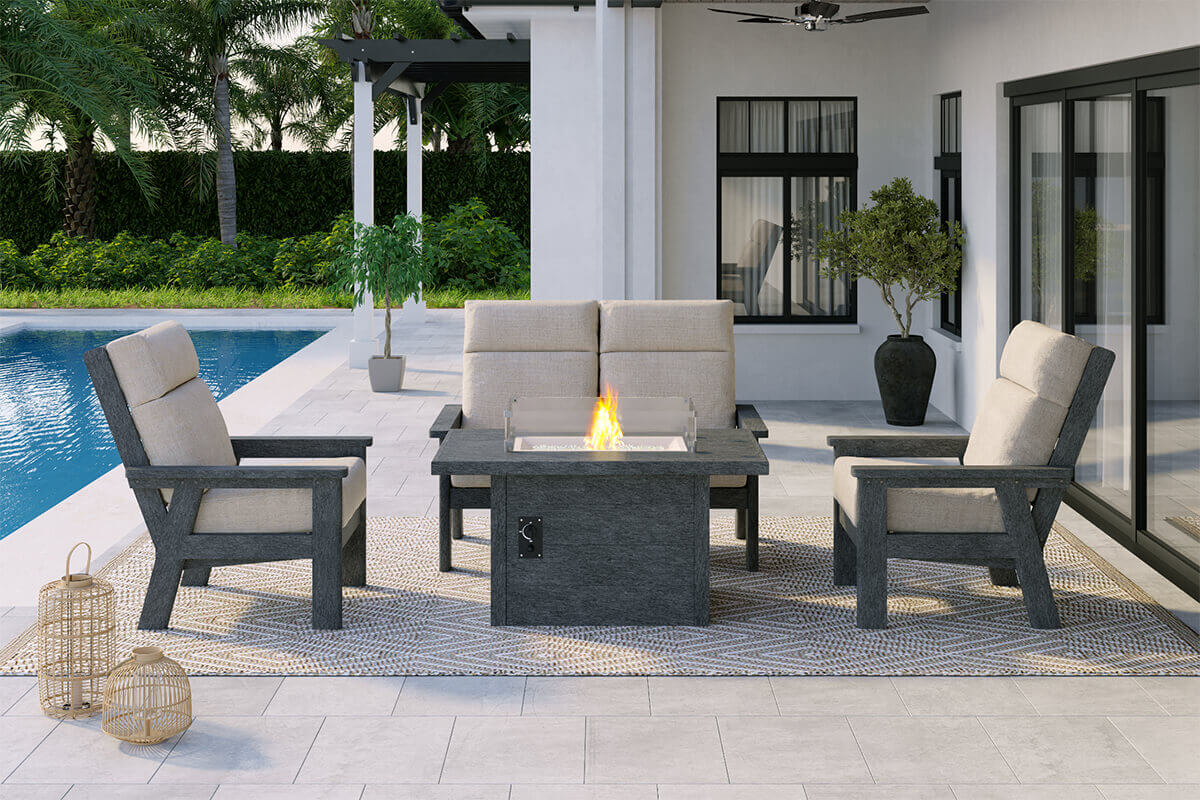 A stylish poolside patio where the CRP rectangular fire table glows with bright flames behind the clear wind guard. Beige cushioned chairs with dark grey recycled plastic frames form a conversation set around the table. The white tiled patio, lush hedges and modern home exterior create a clean and contemporary outdoor living space centred around the 55,000 BTU burner.