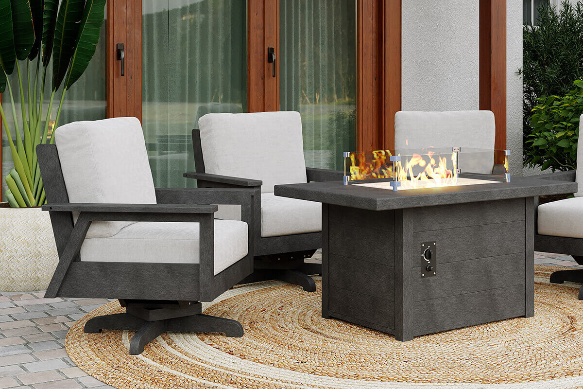 A backyard patio with grey swivel armchairs arranged around the CRP rectangular fire table, which is illuminated with warm flames behind the clear wind guard. The table is built from dark grey recycled plastic lumber with a visible control panel on the side. Pavers, leafy plants and sliding glass doors complete the inviting outdoor entertainment area.