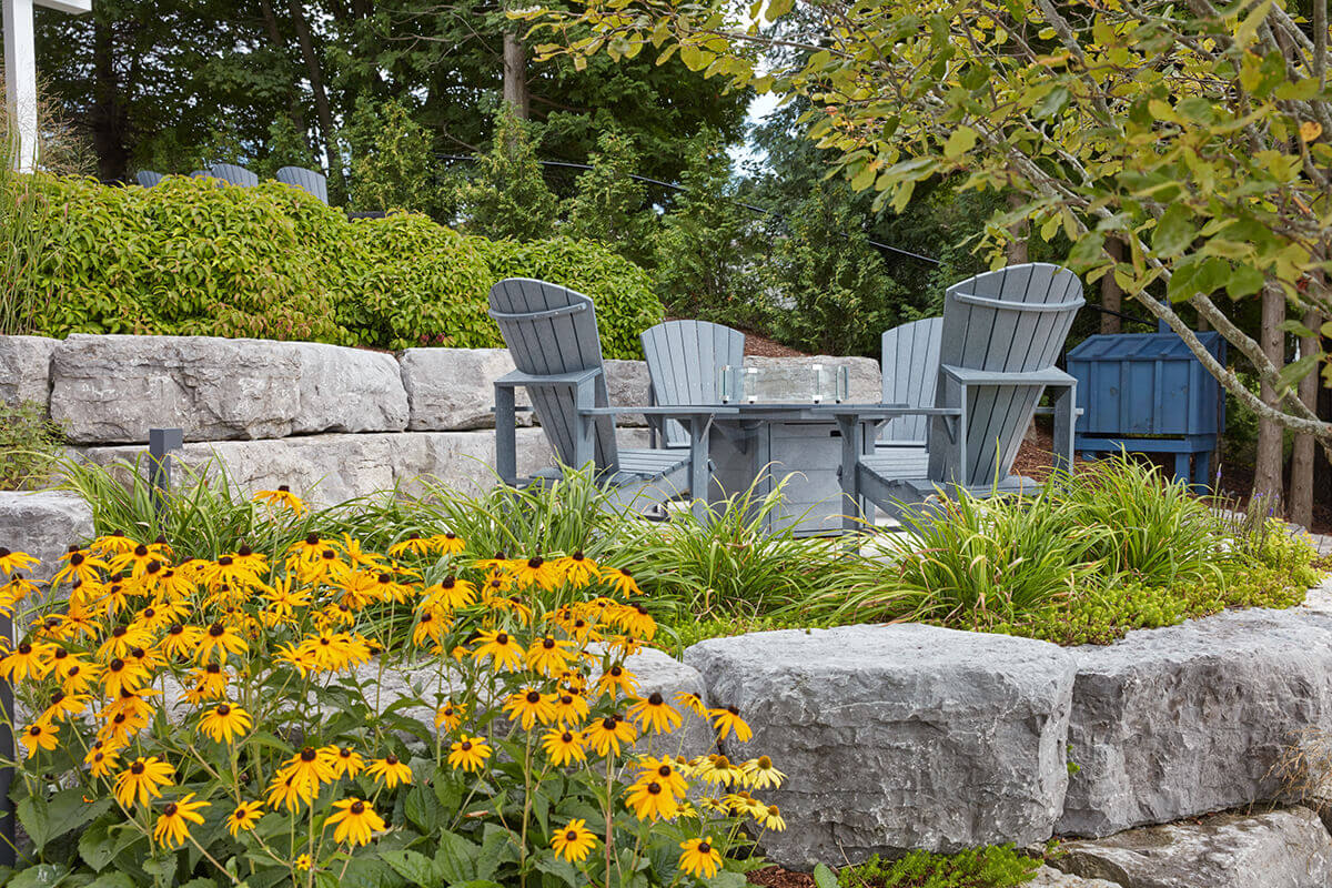 An outdoor seating area featuring a CRP Round Fire Table positioned on a stone patio, surrounded by tall grey recycled-plastic Adirondack chairs. The table has a circular top with a textured grey finish and a raised clear glass wind guard encircling a recessed silver burner tray filled with translucent fire glass beads. Lush landscaping frames the background, including low green shrubs, tall ornamental grasses, large stone retaining walls, and mature trees, creating a layered garden setting. Bright yellow coneflowers with dark centres fill the foreground, contrasting with the cool grey furniture. The scene is lit with soft natural daylight, highlighting the recycled-plastic wood-grain texture of the table and chairs, and reinforcing the fire table’s durable, weatherproof construction.