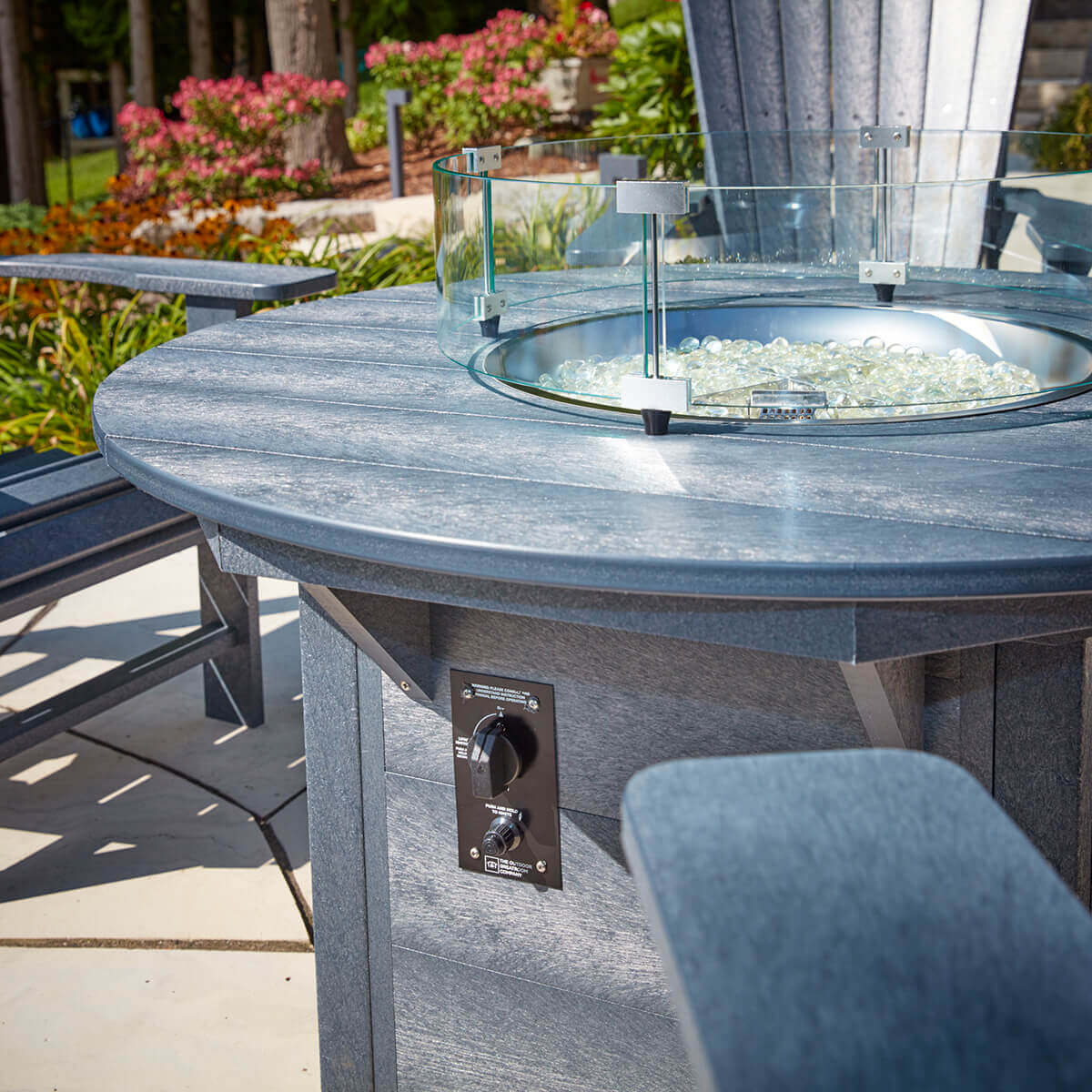 A close angled view of the CRP Round Fire Table’s grey recycled-plastic tabletop showing the circular burner opening surrounded by a curved clear glass wind guard supported by four aluminum corner brackets. Inside the burner tray, dozens of clear fire glass beads catch the sunlight, creating sparkling reflections. Below the tabletop, the ignition control panel is visible on the table’s vertical panel, featuring a black metal faceplate with a rotary flame control knob and push-button ignition. The textured grey recycled-plastic slats reveal subtle grain patterns and reinforcing brackets beneath the tabletop. Tall flowering shrubs and evergreens in the background soften the scene with bright summer colours and dappled shadows.