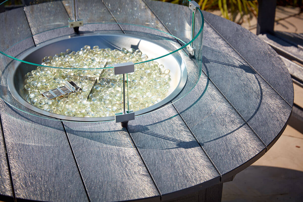 A close view of the CRP Round Fire Table showing its circular tabletop made from textured recycled plastic boards in a deep charcoal colour, with a raised clear glass wind guard forming a full circle around the fire bowl. Inside the bowl sits a bed of pale reflective fire glass that catches the sunlight. Shadows from the wind guard supports fall across the tabletop, revealing the grain-like texture of the material. The focus is on the surface detail and the steel rim surrounding the burner, highlighting the premium outdoor construction.