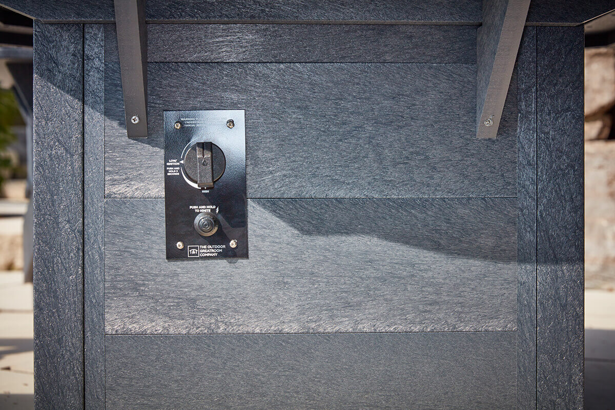 A detailed close up of the control panel on the side of the CRP Round Fire Table, showing a black rectangular metal plate fixed to the charcoal coloured recycled plastic panels. The panel includes a round ignition dial with clear low to high markings, a push to ignite button beneath it and subtle branding from The Outdoor GreatRoom Company. The textured boards around the panel show natural looking grain patterns, emphasising durability for outdoor use.