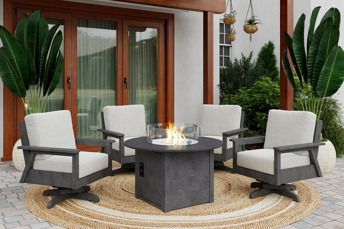 The CRP Round Fire Table positioned on a circular woven rug on a stone patio, surrounded by four cushioned outdoor swivel chairs with grey frames and off white seats. The fire table displays a lively flame rising from the round burner encircled by a clear wind guard, casting a warm glow across the chairs. Behind the setup is a modern home with wooden framed sliding glass doors and tall potted tropical plants, creating a relaxing outdoor living mood.