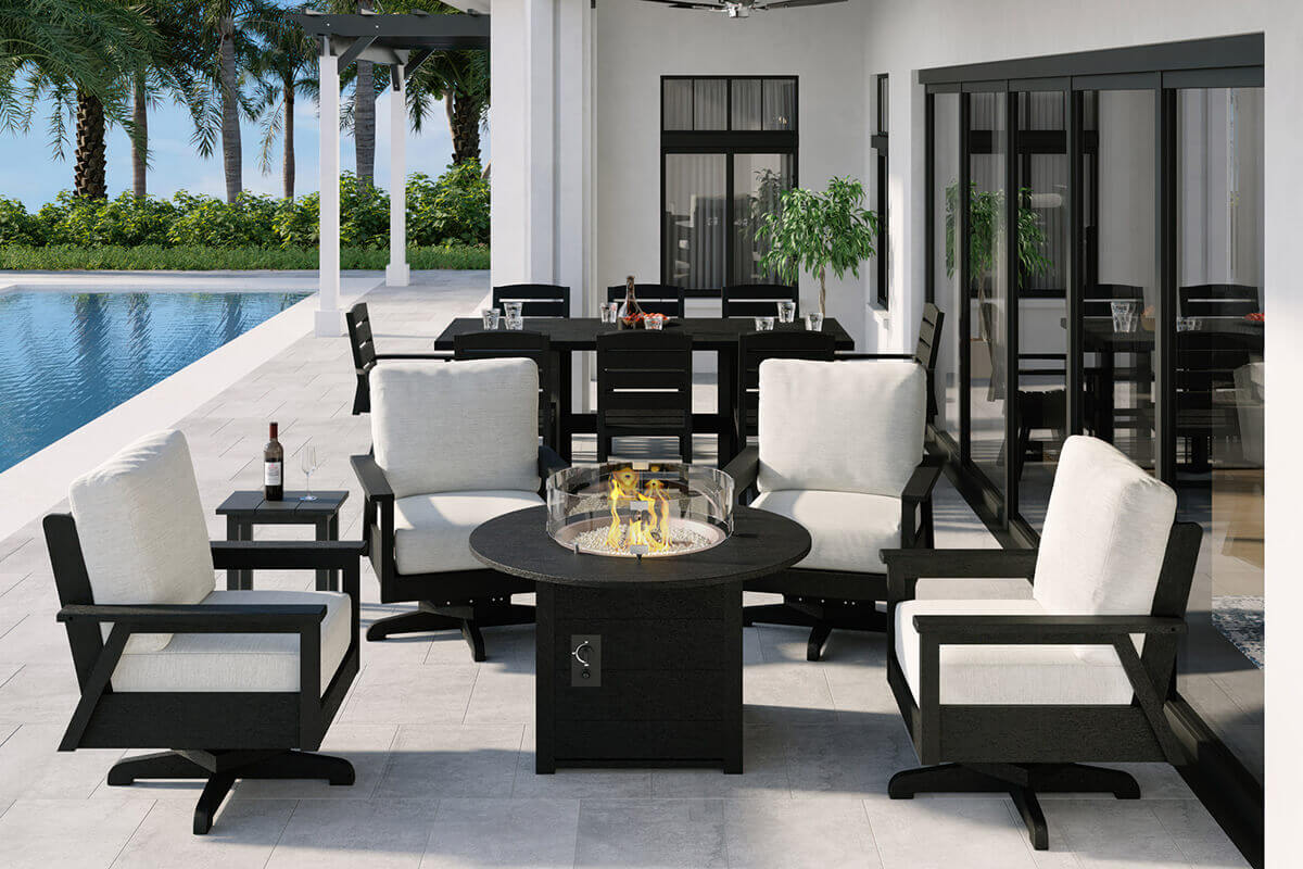 A poolside setting featuring the CRP Round Fire Table centred among four matching outdoor swivel chairs with off white cushions and black frames. The fire table emits a tall warm flame inside the circular burner, protected by a clear wind guard. The surrounding patio is modern and clean with a dining set in the background and a lush line of palm trees forming a privacy wall. The fire table stands out as the glowing focal point of the seating area.