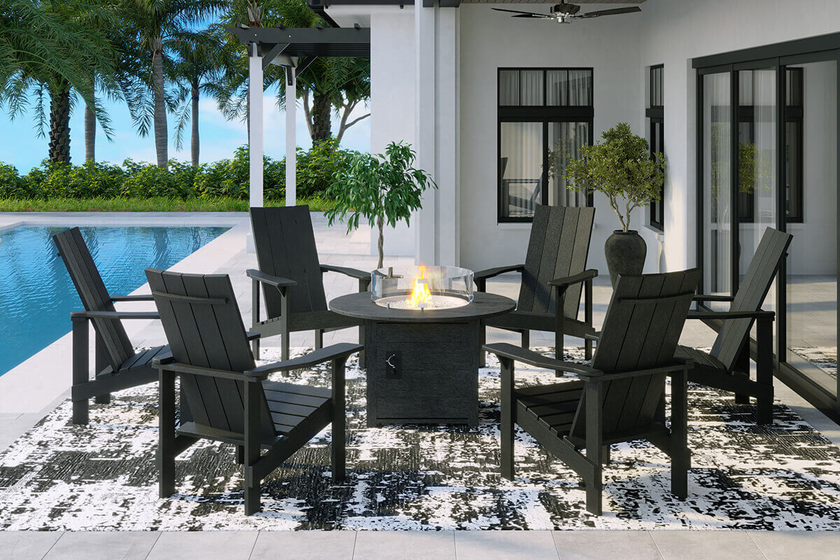A modern patio with the CRP Round Fire Table surrounded by Adirondack style black outdoor chairs arranged in a wide circle. The fire table’s round charcoal top supports a glass wind guard containing a vibrant flame. The patio sits beside a bright blue pool with palm trees and a pergola in the background. The contrast between the dark furniture and the glowing flame under bright daylight creates a welcoming social space.