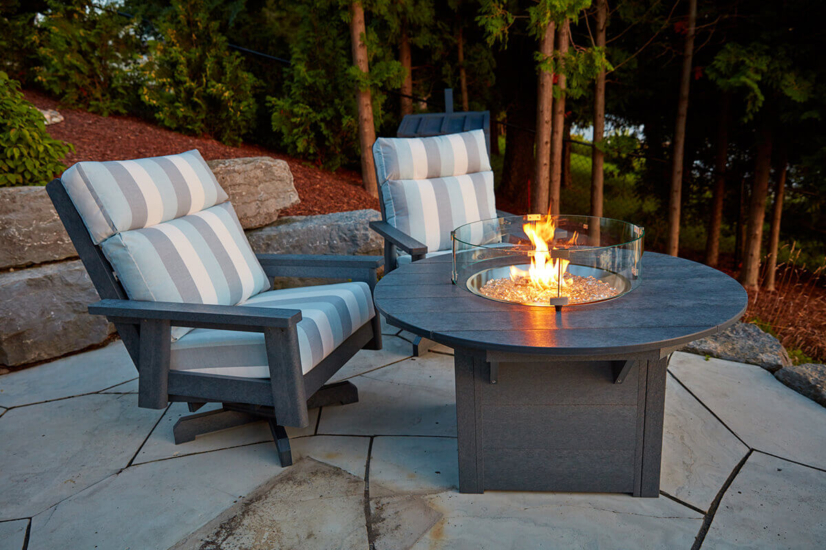 Two cushioned outdoor chairs with wide armrests face each other beside the CRP Round Fire Table on a paved lakeside patio. The fire table glows with an active flame inside the clear wind guard, reflecting off the glass and fire media. The scene is framed by rocky landscaping and tall evergreens as the sun sets, casting soft shadows and creating a peaceful waterside ambience.