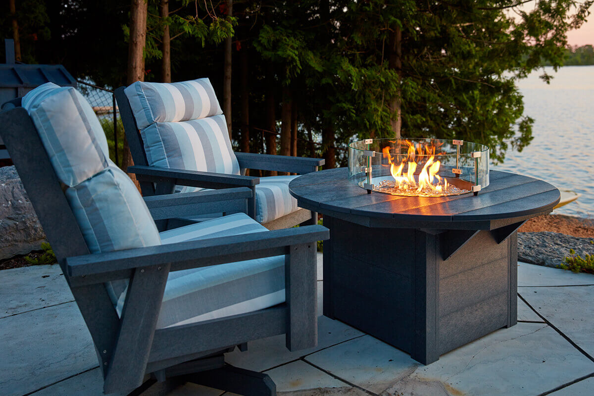 A view of the CRP Round Fire Table from behind two cushioned outdoor chairs angled toward a lake at sunset. The flame rises strongly within the circular burner, surrounded by the transparent wind guard. Warm amber light spreads across the charcoal textured tabletop as the lake reflects soft evening colours. This lakeside perspective highlights the table as the central gathering point for quiet outdoor relaxation.