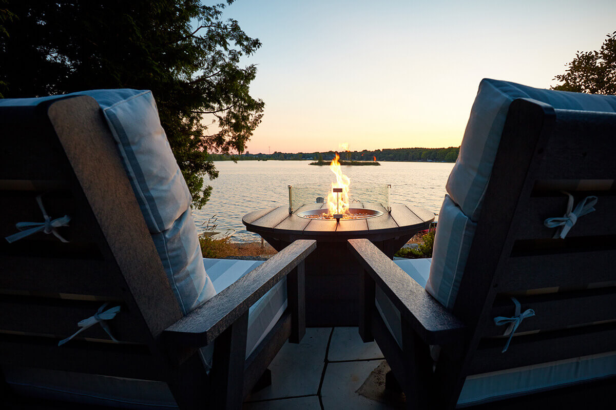 A twilight scene showing the CRP Round Fire Table glowing beside two striped cushioned outdoor chairs positioned on a stone patio. The round tabletop displays its textured charcoal boards clearly illuminated by the flame inside the circular burner. Behind the chairs stand tall trees and a sloped landscaped garden, while the lake reflects the fading sunset. The warm firelight contrasts with the cool outdoor colours, enhancing the inviting atmosphere.