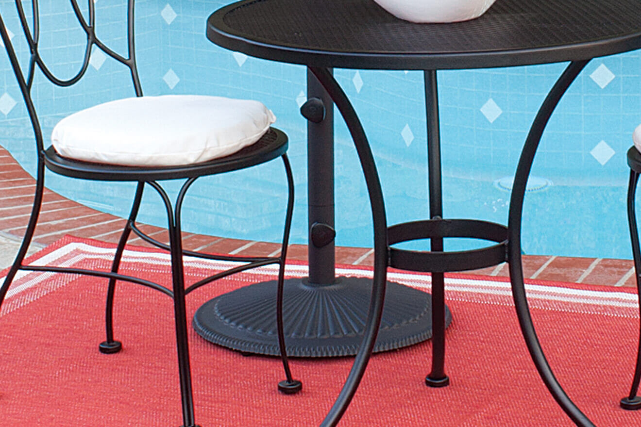 Outdoor close-up of a round black Classic Cast Iron Umbrella Base placed beneath a matching wrought iron patio table and chair set beside a swimming pool. The umbrella pole rises from the textured cast iron base, which features a decorative radial pattern. The black powder-coated surface contrasts against the red outdoor rug and blue water tiles in the background, highlighting its elegant yet sturdy design that complements the furniture ensemble.