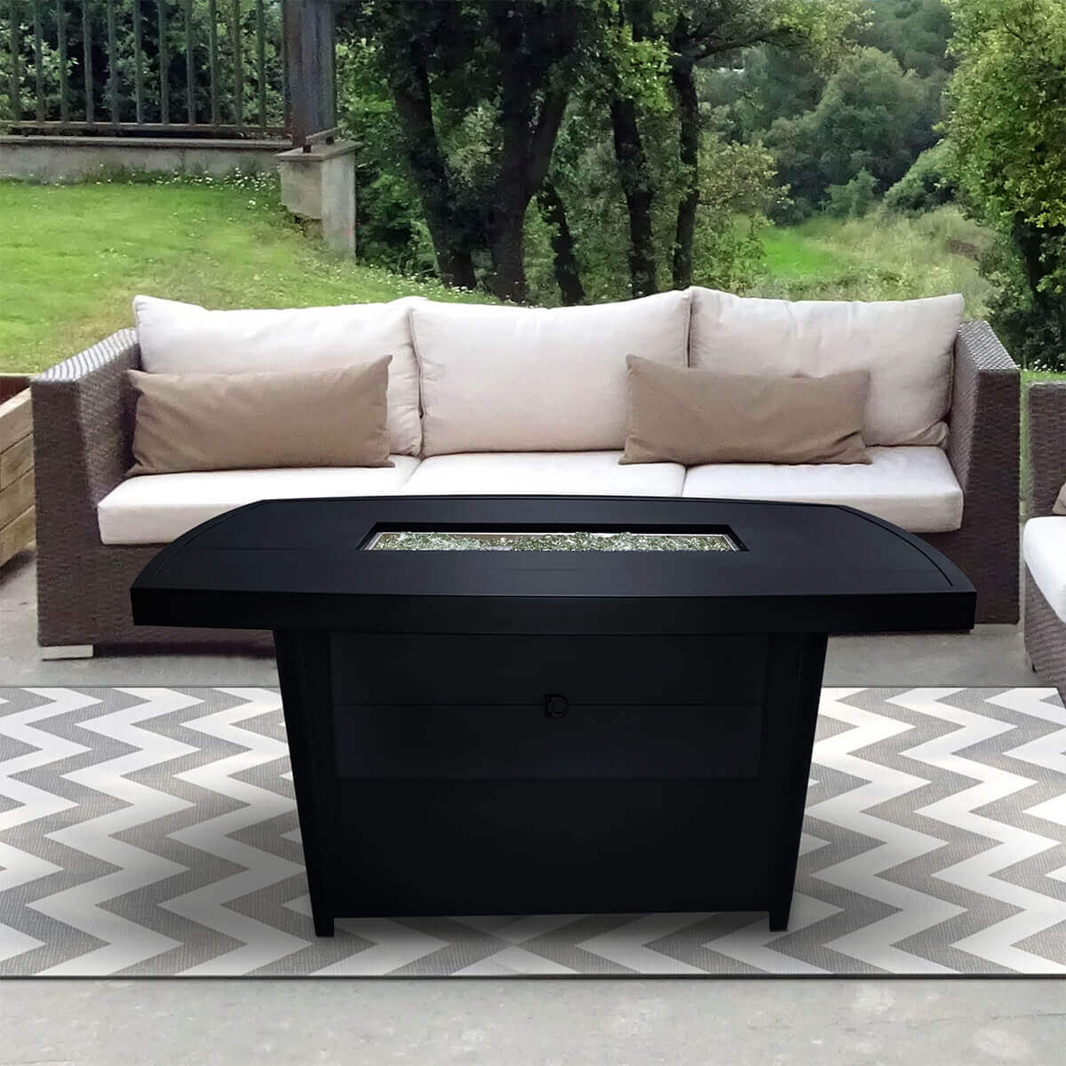Highly detailed view of the Sunbeam Edge Fire Table shown outdoors on a grey and white chevron rug in front of a beige outdoor sofa. The rectangular aluminum fire table in a smooth black onyx finish sits centred in the frame, with its closed burner area visible as a recessed rectangle filled with clear kidney shaped glass beads. The wide curved tabletop tapers elegantly toward the middle, while the lower cabinet features a flush surface with a circular cutout for the concealed propane door near the bottom centre. The table’s shadow falls softly onto the rug, and lush green trees form the natural backdrop, enhancing the product’s contrast and presence. SEO keywords included: Sunbeam Edge Fire Table, black onyx aluminum fire table, outdoor propane fire pit.