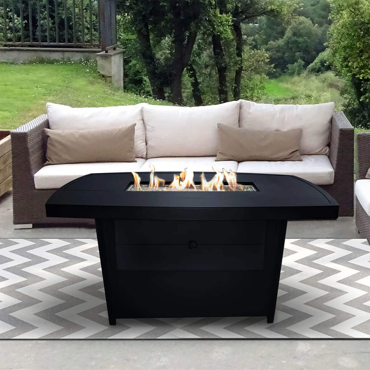 Sunbeam Edge Fire Table displayed outdoors with its burner ignited, creating vivid dancing flames rising from the rectangular recessed burner tray filled with shimmering kidney shaped glass beads. The black onyx aluminum frame contrasts sharply with the glowing fire, and the flame reflection softens across the smooth tabletop surface. The table stands on a chevron patterned rug between two wicker sofas with cream cushions. Behind the setup, tall green trees and open grass create a serene natural backdrop, highlighting the warm evening ambience. SEO keywords included: Edge Fire Table with flames, outdoor aluminum fire pit, Sunbeam fire table lit.