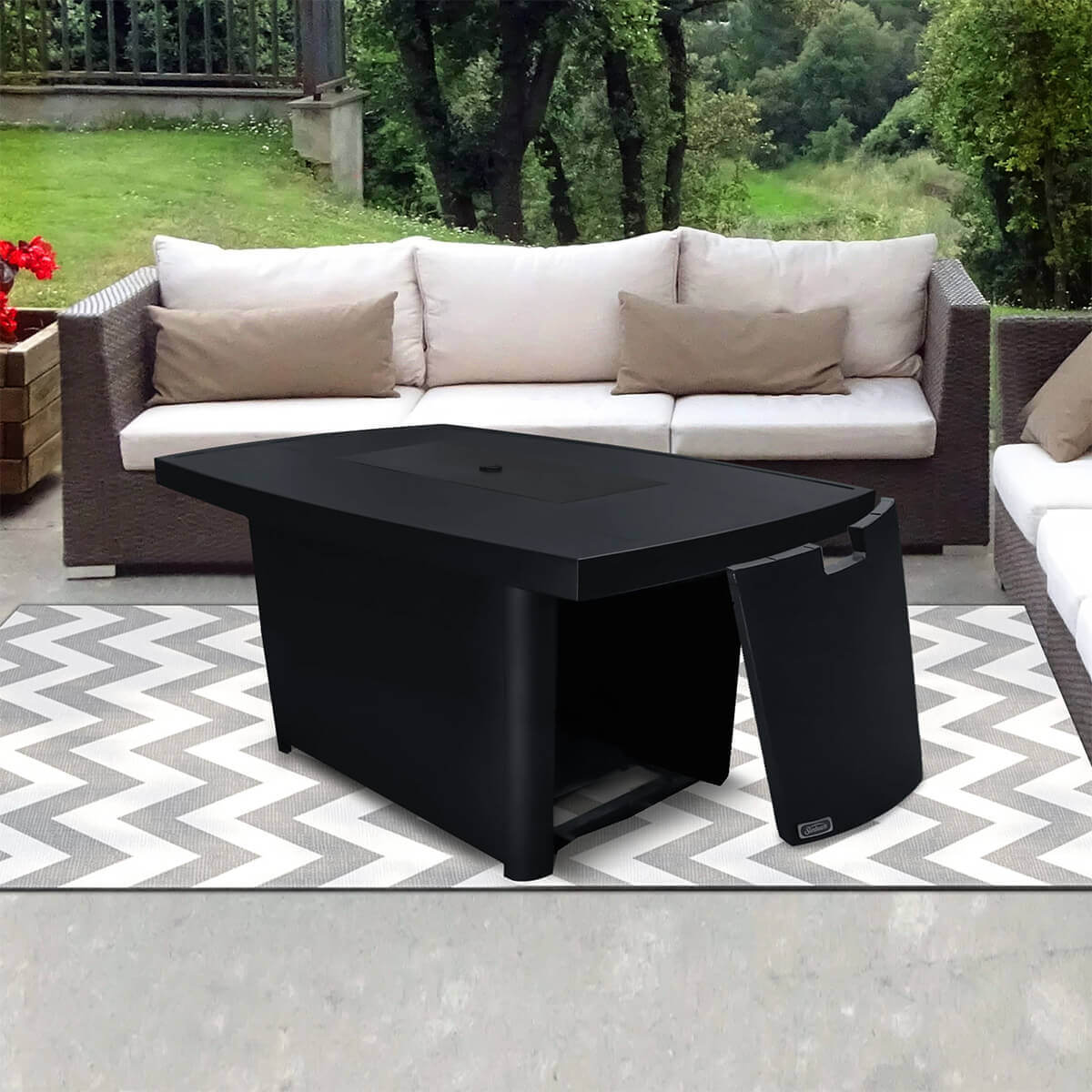 Side angled view of the Sunbeam Edge Fire Table with the metal lid placed on top, fully covering the burner area to convert the unit into a functional table surface. The black onyx aluminum lid sits flush within the rectangular opening, maintaining clean uninterrupted lines across the tabletop. To the right of the table, the easy access propane door is shown removed and placed leaning upright on the ground, revealing the interior storage area. The table stands on a chevron patterned rug surrounded by beige outdoor sofas, with lush trees and grass in the background. SEO keywords included: Edge Fire Table with lid, aluminum fire table with propane access, Sunbeam outdoor table.