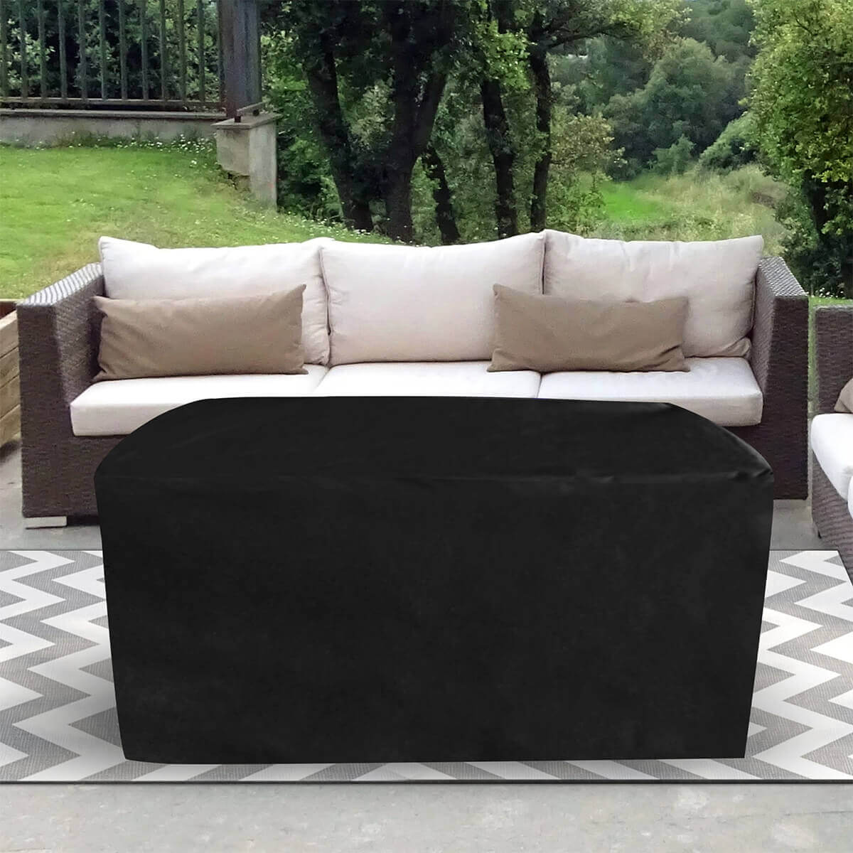 The Sunbeam Edge Fire Table fully covered with its fitted black protective cover, draped smoothly across the entire rectangular silhouette. The cover extends down all sides, hiding the aluminum body and giving the appearance of a seamless block shape. The table sits on a chevron patterned outdoor rug between beige cushioned sofas, with a backdrop of green trees and raised garden features. No burner components or structural details are visible due to the full coverage. SEO keywords included: Edge Fire Table cover, outdoor fire table protection, Sunbeam fire pit cover.