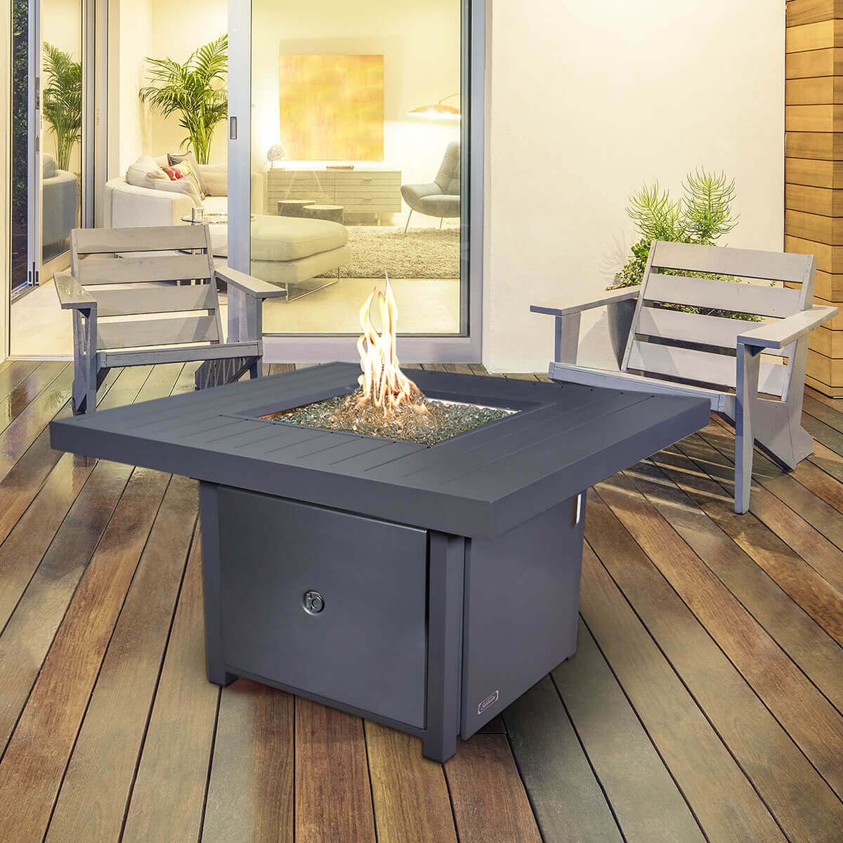 A warm grey Sunbeam Fabia Fire Table displayed on a modern wooden deck, shown with its square aluminum tabletop surrounding a recessed fire pit filled with shimmering kidney shaped glass beads. Bright golden flames rise from the centre, contained within a stainless steel burner. Two contemporary grey patio chairs sit on either side, positioned in front of large sliding glass doors revealing a softly lit living room. The table’s broad slatted aluminium top, clean modern lines and sturdy base create a sophisticated focal point, emphasising its adjustable flame feature, easy push button ignition and outdoor luxury aesthetic.