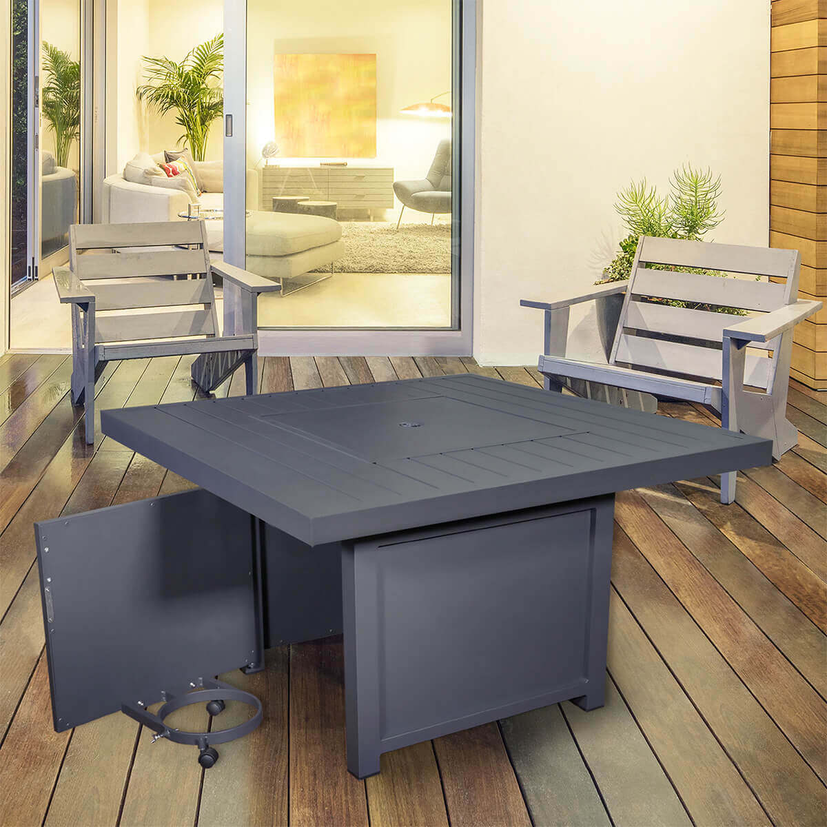 The Sunbeam Fabia Fire Table shown with the metal lid securely covering the fire bowl, creating a fully flat square surface. The hinged propane access door is open, revealing the circular swivel tray designed for holding a 20 lb propane tank. The warm grey aluminum structure has clean vertical lines and a smooth slatted tabletop. Two modern grey patio chairs flank the table, and the background features a glowing indoor living space visible through large sliding doors, reinforcing the fire table’s versatility and elegant outdoor integration.
